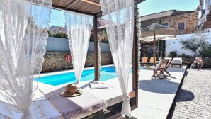 A covered outdoor seating area with a pool, stone walls, and a rustic building in the background.
