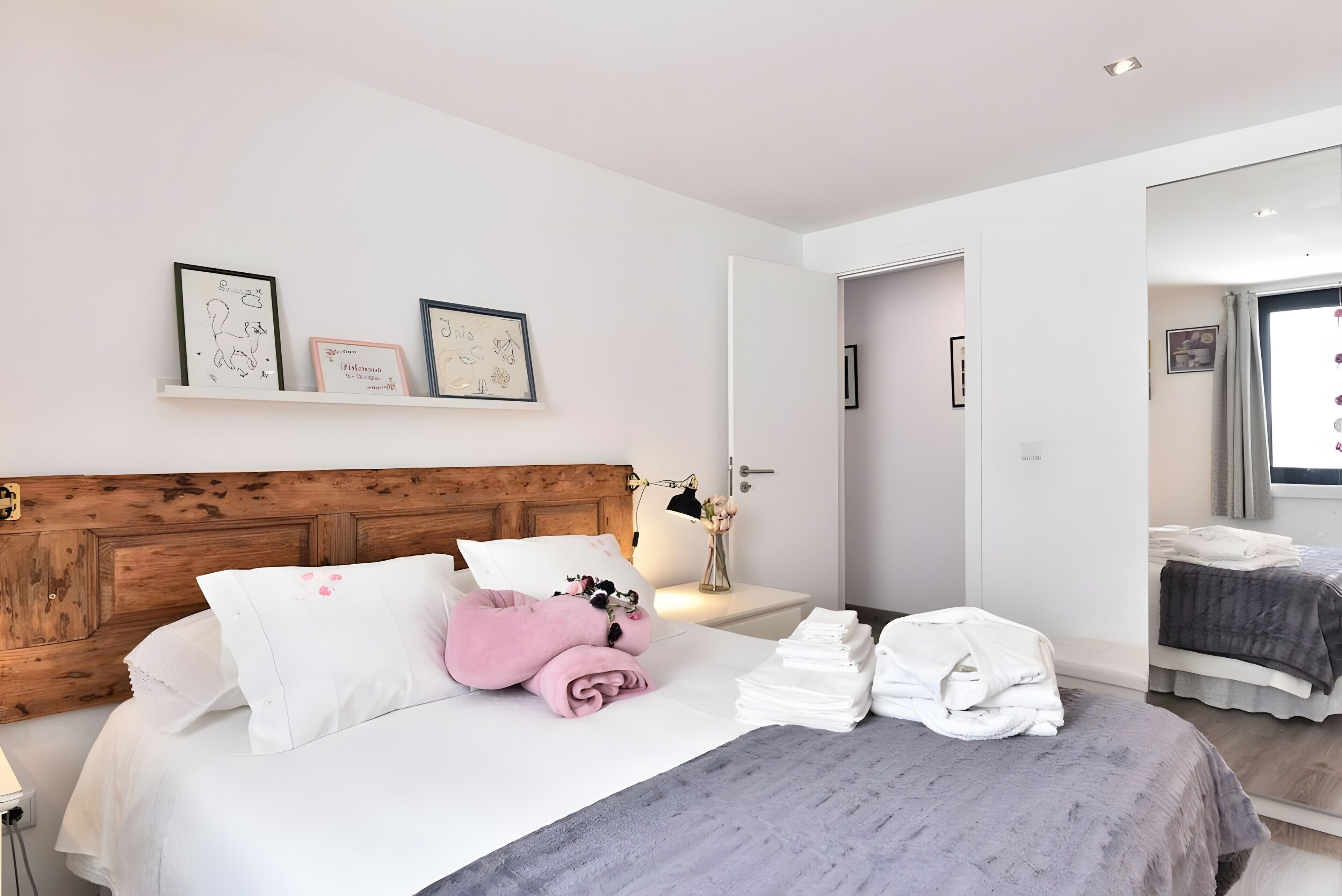 Habitación de hotel con cabecera de madera, cama con ropa de cama blanca y rosa, mesita de noche con lámpara y un jarrón, y vista a otra habitación a través de una puerta abierta.