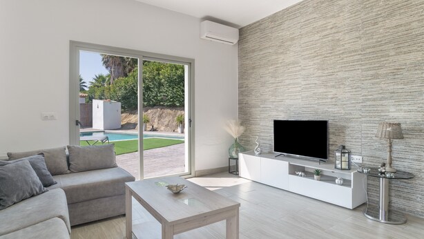 En moderne stue med et fladskærms-tv, en sofa, et sofabord og en stor skydedør, der fører til et udendørs område med en pool.