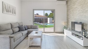 En moderne stue med en grå sofa, et hvidt sofabord, et fjernsyn på et hvidt skab og en skydedør, der fører til en pool.