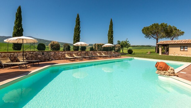 A swimming pool with lounge chairs and umbrellas, surrounded by stone walls and tall trees.