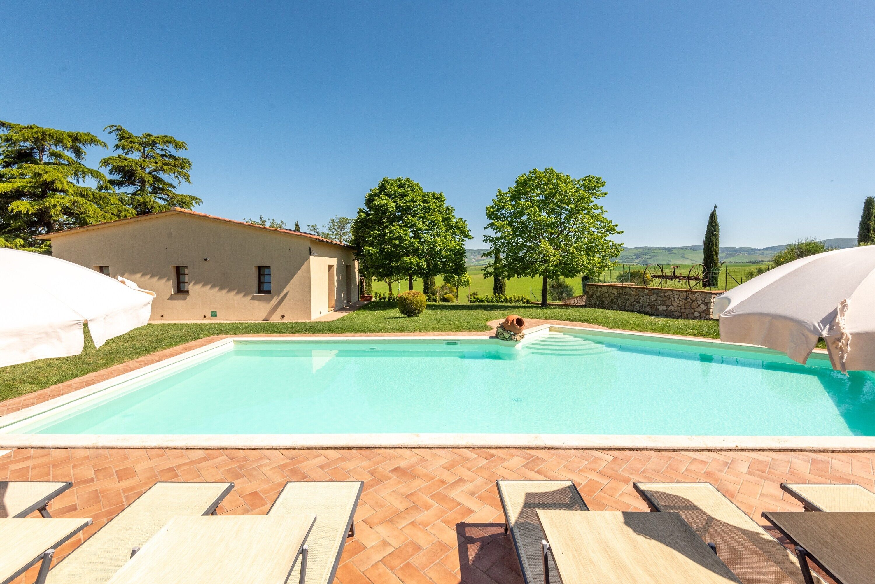 Una piscina con lettini e ombrelloni, circondata da un edificio e alberi.