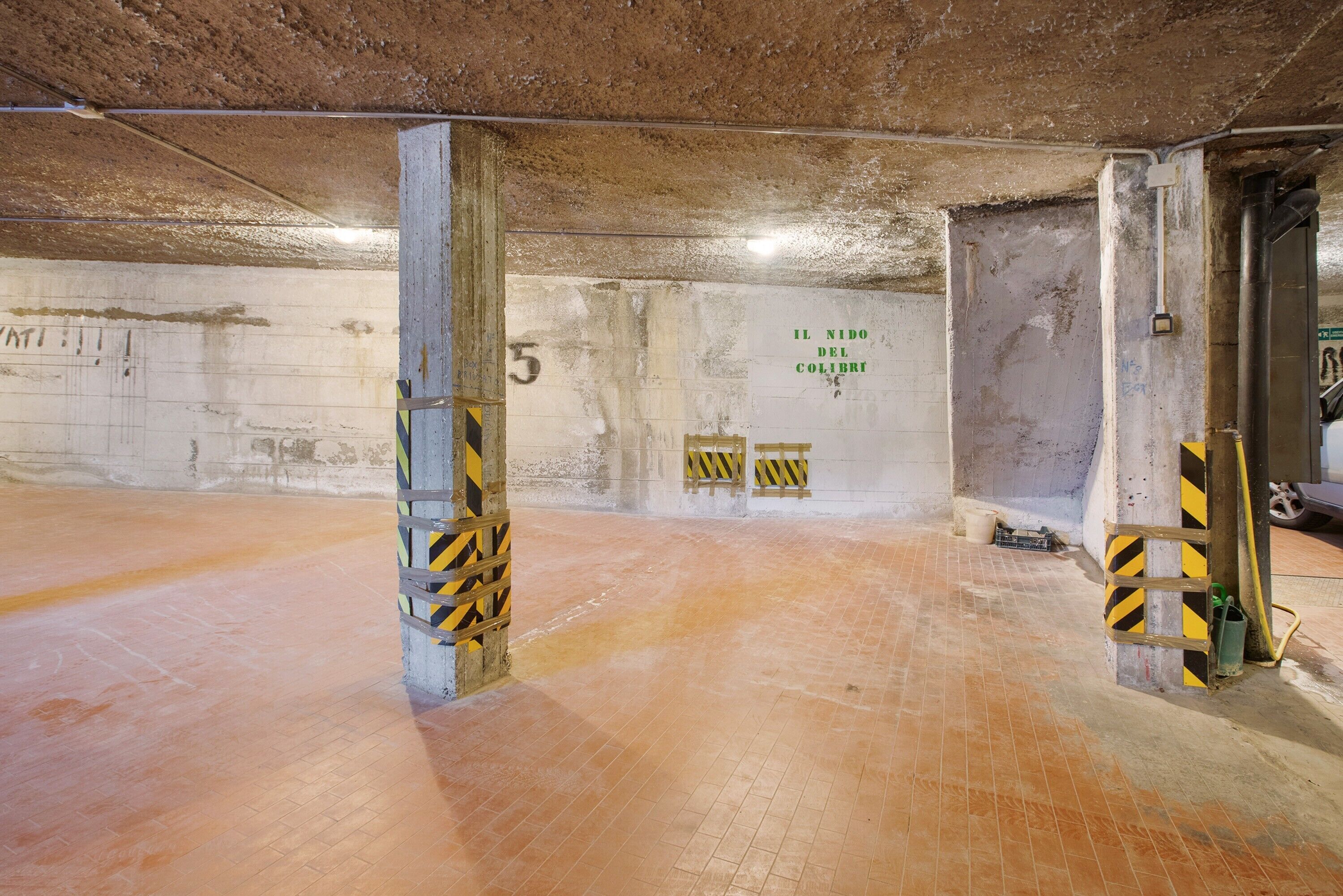 An underground parking area with concrete pillars, a tiled floor, and a wall with graffiti.