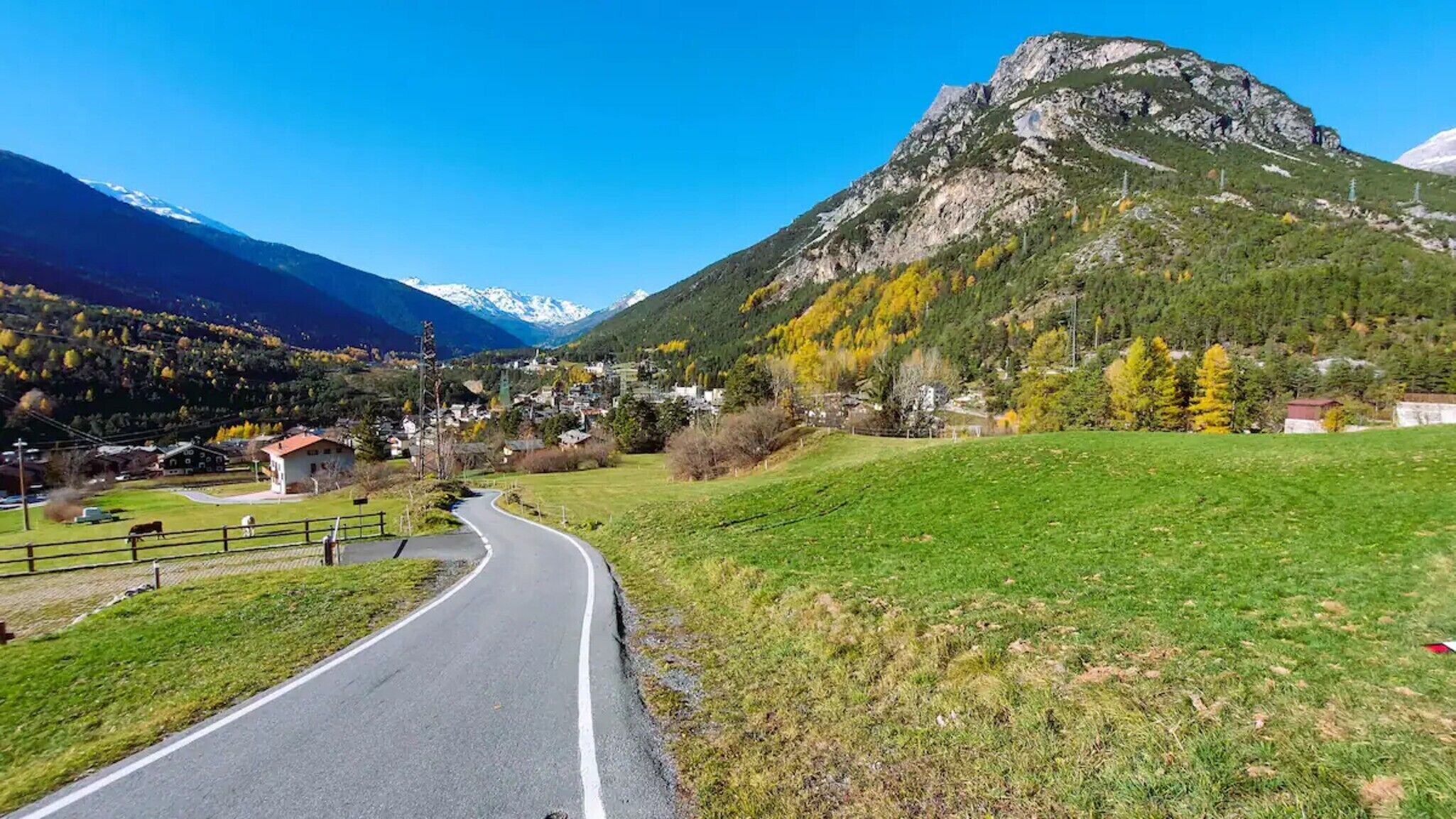A rural road curving through a valley with a mountain backdrop, houses, and a grassy field.
