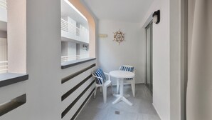 Un balcón estrecho con una mesa y sillas blancas, un adorno en la pared y vistas del interior del edificio.