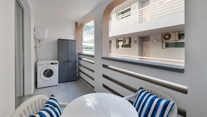 Un balcón con lavadora, una mesa con cojines a rayas y vistas a un edificio con balcones.