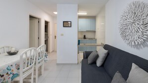 Una sala de estar moderna con un sofá gris oscuro, una mesa de comedor blanca con sillas y una cocina al fondo.