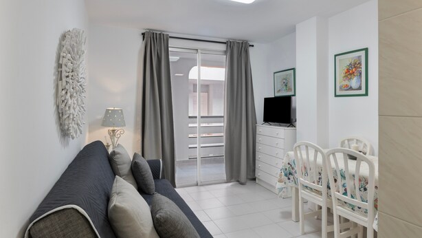 Una sala moderna con sofá, una mesa de comedor con sillas, televisor y un balcón con cortinas.