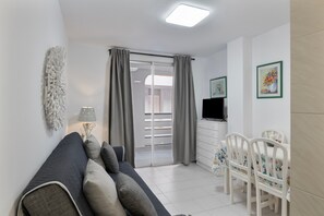 A modern living room with a sofa, a dining table with chairs, a TV, and a balcony with curtains.