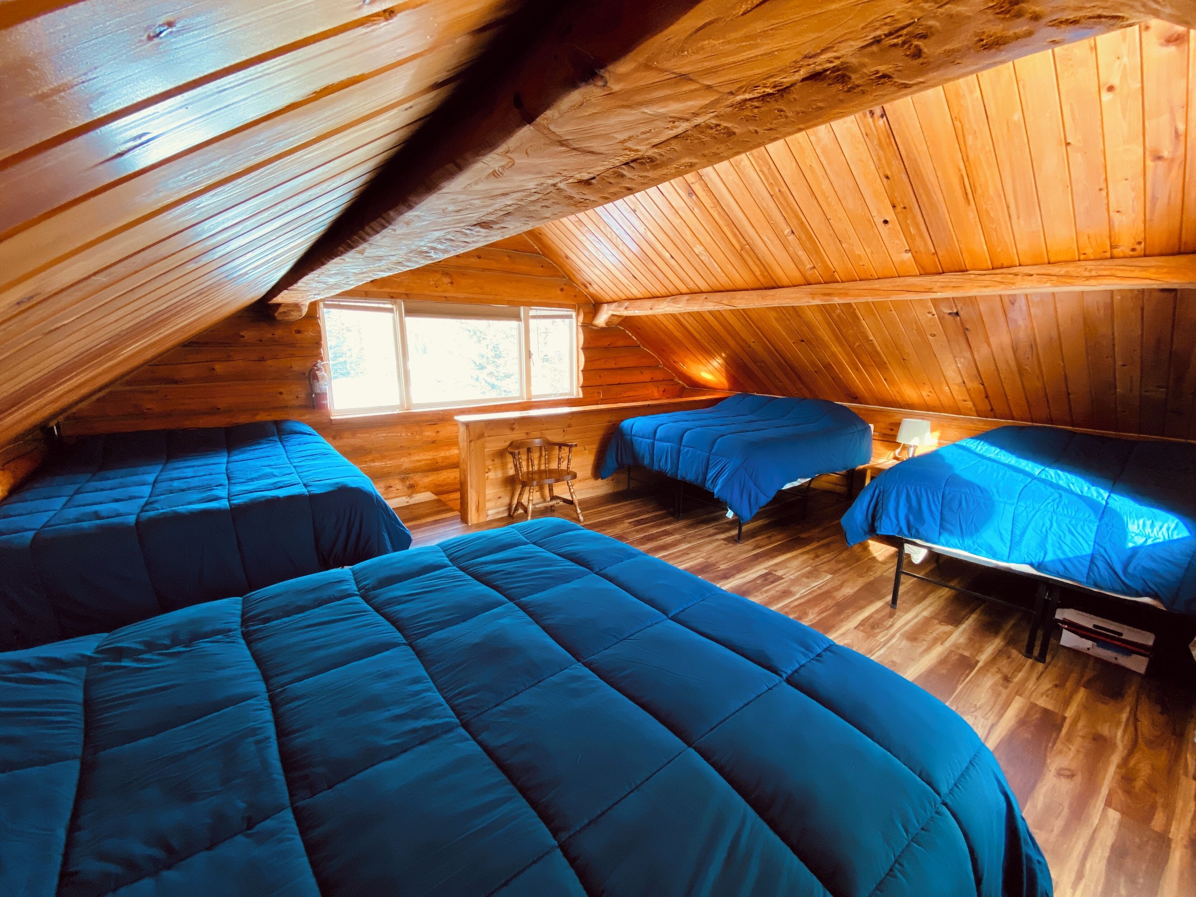 A cozy attic bedroom with three beds, wooden walls, and a window providing natural light.