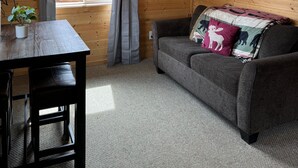 L’intérieur d’un chalet accueillant, avec un mur en bois, un canapé gris agrémenté de coussins à motifs animaliers, une petite table sur laquelle est posée une plante en pot, et une fenêtre avec des rideaux.