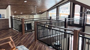 Ett rymligt rum i loftstil med högt tak, trägolv, ett svart metallräcke och utsikt över ett snötäckt landskap genom stora fönster.