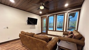 A living room with a brown sofa, a small table, and a TV mounted on the wall. There are large windows with a view of a forest and a lake.