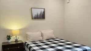 Ett sovrum med ett rutigt sängöverkast, ett nattduksbord med en lampa och en inramad tavla på väggen.