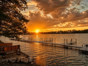 Vista de la puesta de sol sobre un lago, con un muelle, una banca y una fila de árboles.