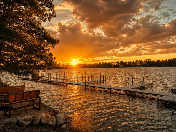 Vista de la puesta de sol sobre un lago, con un muelle, una banca y una fila de árboles.