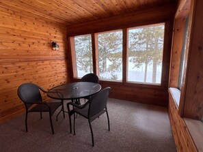 Een chaletinterieur met een ronde tafel en vier stoelen, houten wanden en grote ramen met uitzicht op sneeuw.
