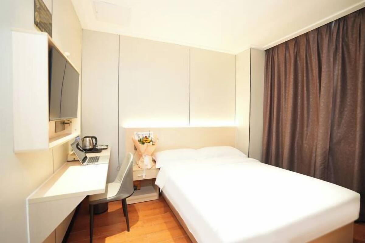 Une chambre d’hôtel avec un lit, un bureau avec un ordinateur portable, une chaise, une petite table avec un vase de fleurs et un téléviseur fixé au mur.