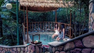 Un área de piscina con paredes de piedra y zona de estar cubierta por un techo de paja, rodeada de abundante vegetación.