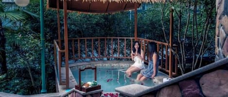 Un área de piscina con paredes de piedra y zona de estar cubierta por un techo de paja, rodeada de abundante vegetación.