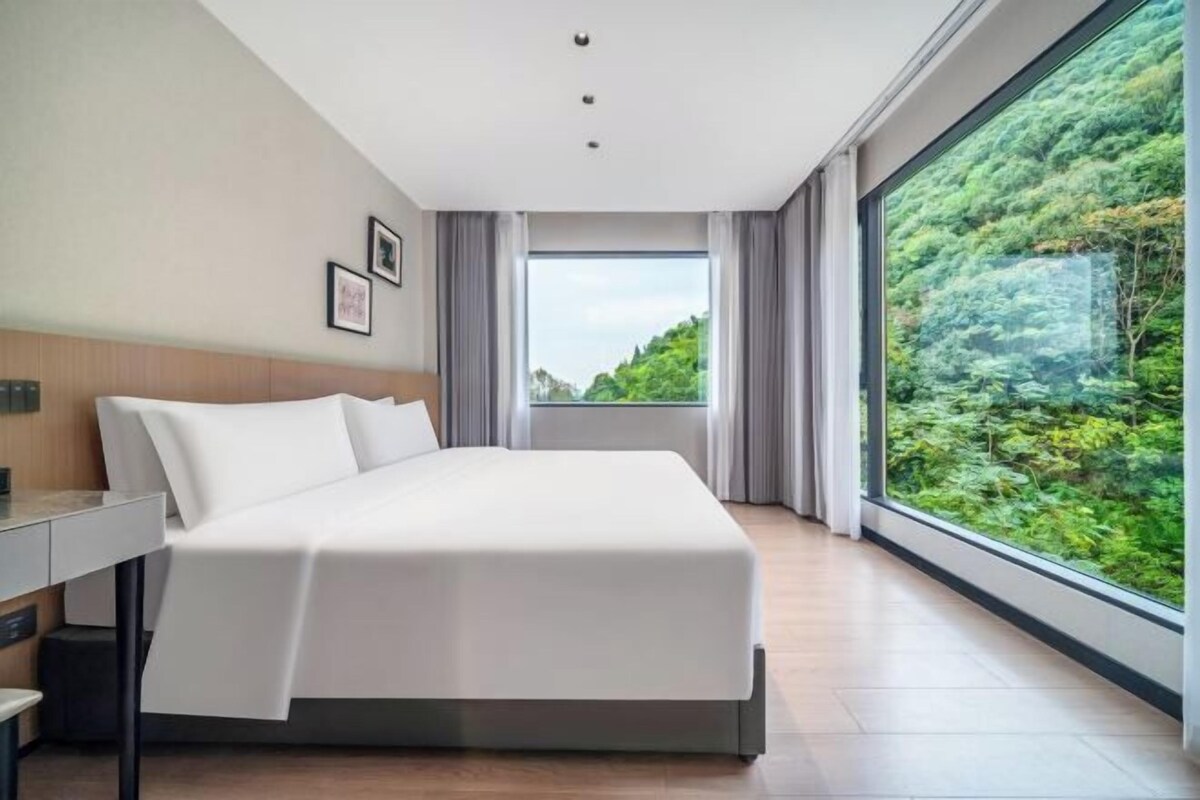 A modern hotel room with a large bed, a desk, and a view of lush greenery through large windows.
