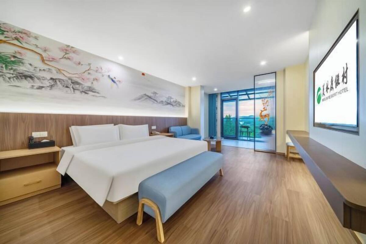 Quarto de hotel moderno com uma cama grande, um banco de madeira, uma escrivaninha e vista para o exterior por uma porta de vidro.