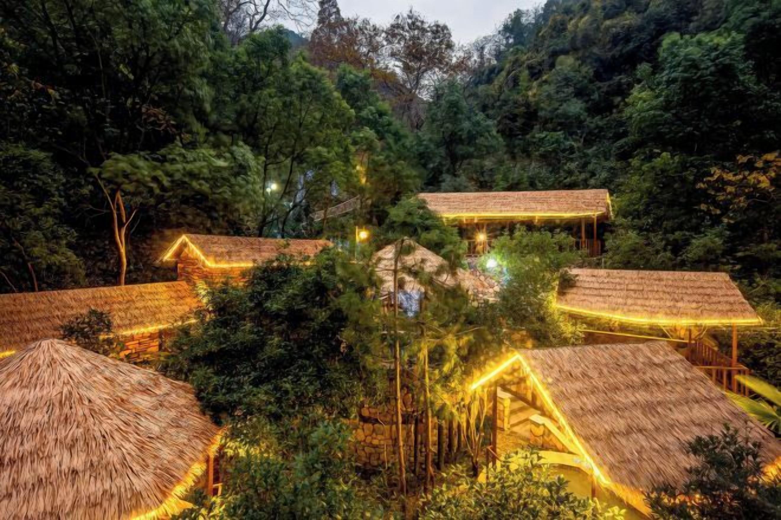 Conjunto de construcciones con techo de paja e iluminación cálida, rodeado de un bosque frondoso.