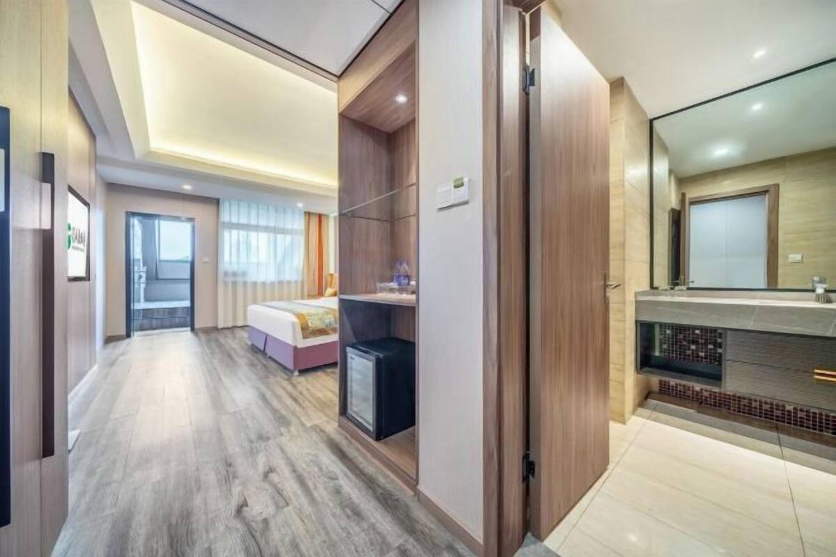 Quarto de hotel moderno com piso de madeira, cama, escrivaninha e banheiro com espelho e pia.