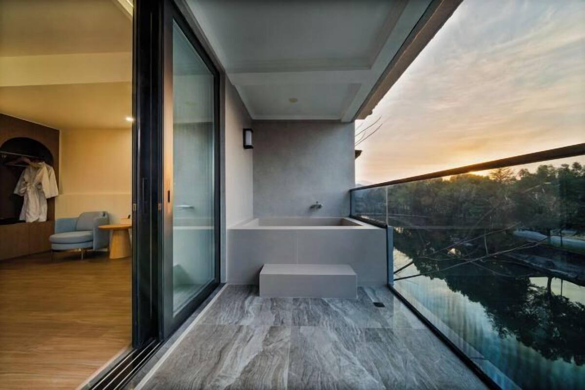 Uma casa de banho moderna com banheira, cabine de chuveiro em vidro e varanda com vista para uma ponte e árvores.