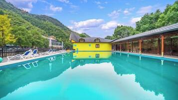 A clear swimming pool with lounge chairs, a yellow building, and a mountain backdrop.