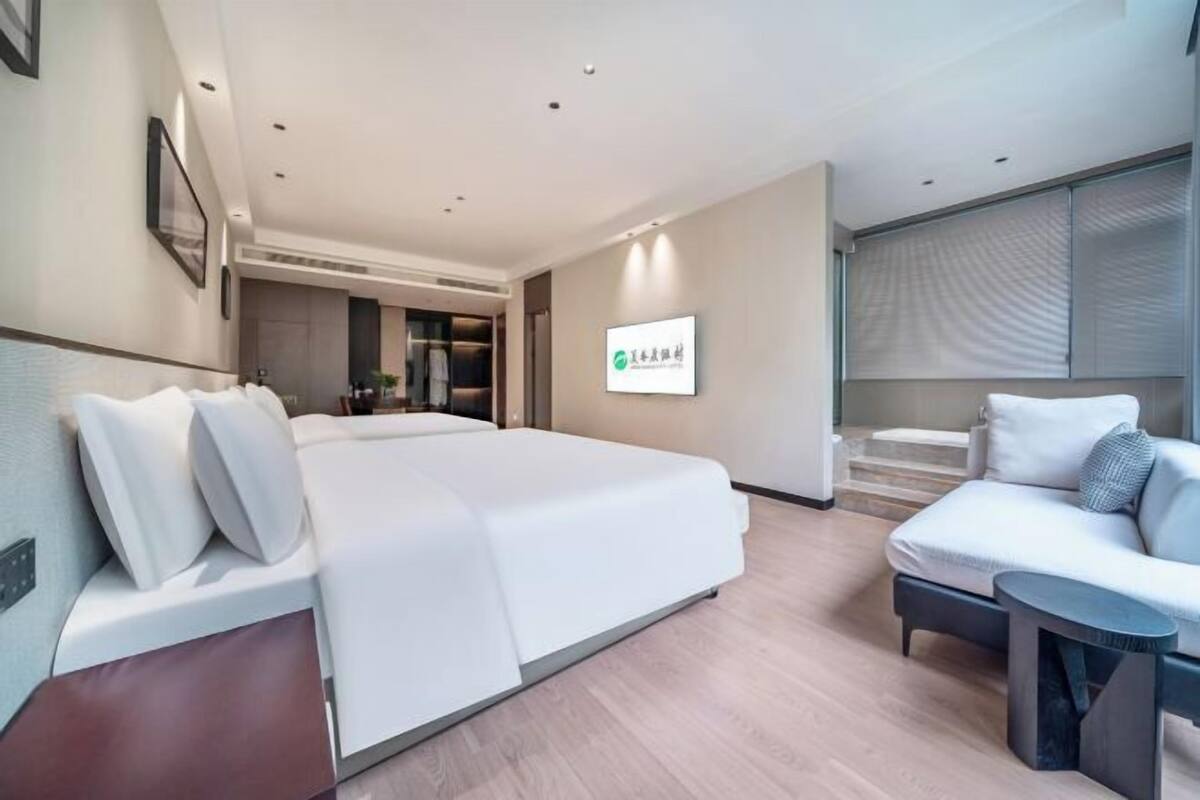 Quarto de hotel moderno com uma cama grande, um sofá, uma mesinha e um banheiro com porta de vidro.