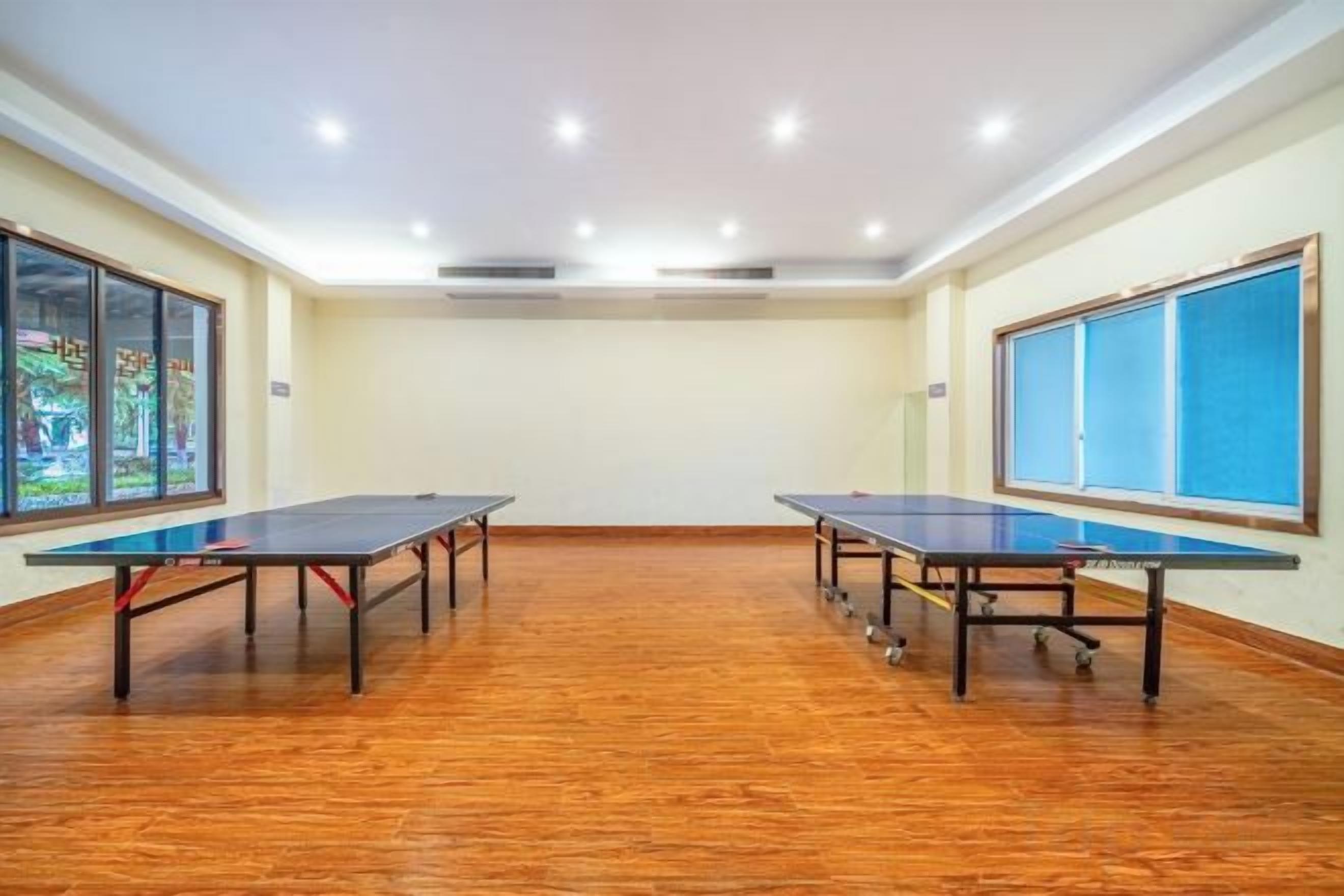A room with two ping pong tables, wooden flooring, and large windows with blue curtains.