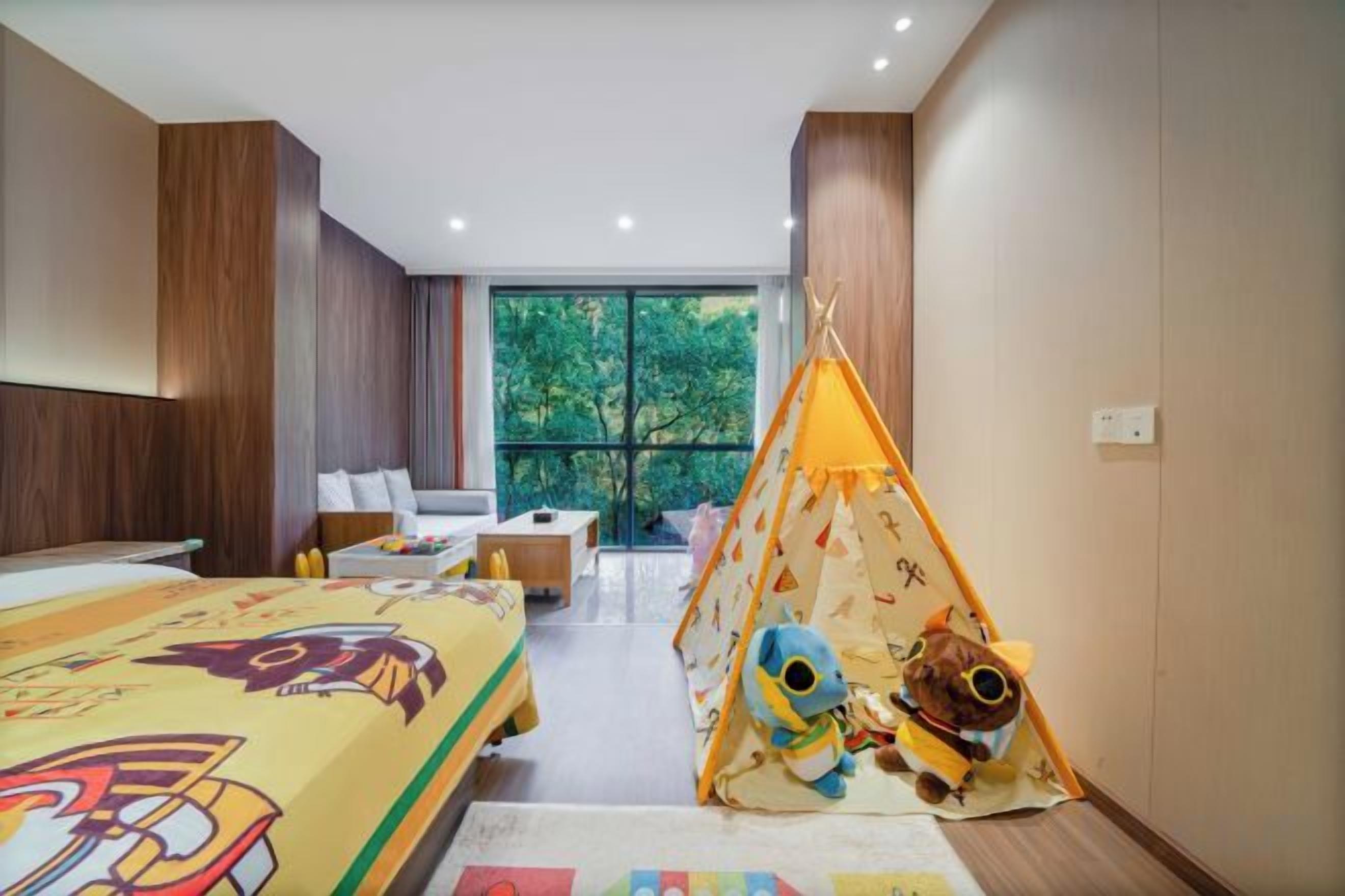 A hotel room with a large bed, a small sofa, a tent with stuffed animals, and a view of trees through the window.
