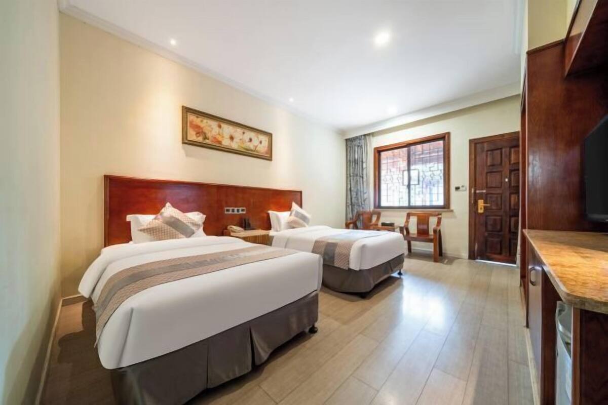 Quarto de hotel com duas camas, cabeceira de madeira, um quadro na parede, janela com cortinas e uma área de cozinha com bancada e armários.