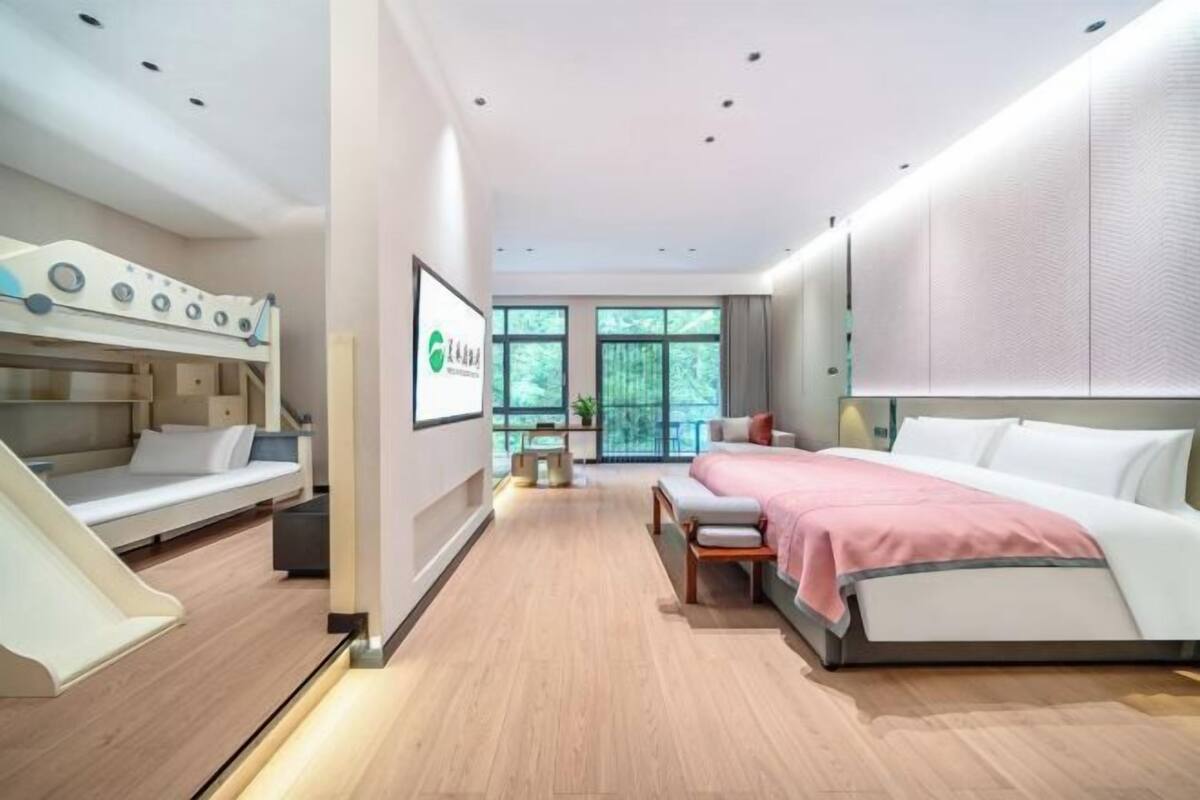 Quarto de hotel moderno com beliche, uma cama grande, escrivaninha e janela com vista para vegetação.