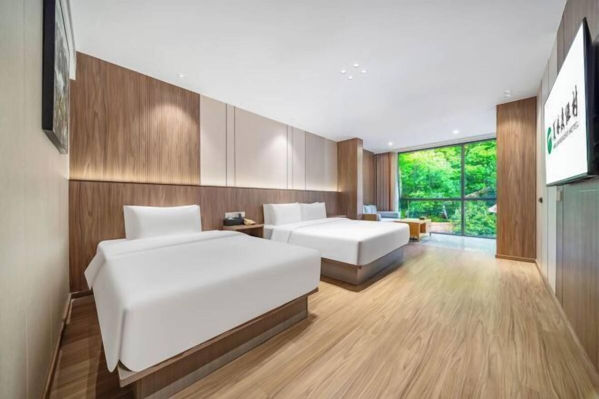 Quarto de hotel com duas camas, painéis de madeira, uma janela grande com vista para vegetação e uma televisão de tela plana.