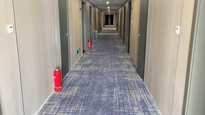 Un pasillo largo y estrecho con puertas a ambos lados, un extintor a la izquierda y una manguera contra incendios a la derecha.