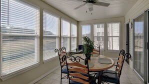 A sunroom with a ceiling fan, large windows, a dining table with chairs, and a potted plant.