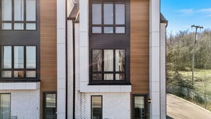 A modern two-story residential building with a combination of white brick and wooden panel facades, large windows, and a visible air conditioning unit. - Cozy Haven in Hermitage (Nashville)