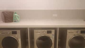 A laundry area with three washing machines and a shelf with a basket and a bottle.