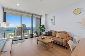 Un salon moderne avec un canapé en cuir, une table basse en bois et une vue sur l’océan et le paysage urbain, grâce à de grandes fenêtres.