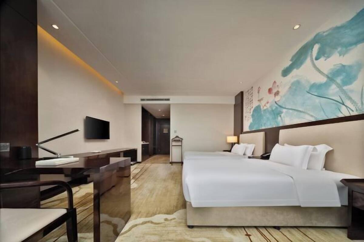 A hotel room with a bed, desk, chair, TV, and artwork on the wall.