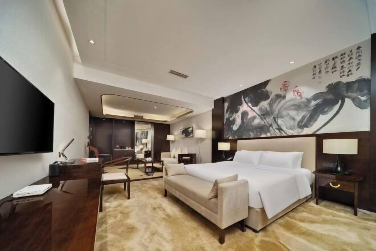 A hotel room with a large bed, a desk, a chair, a TV, and a painting on the wall.