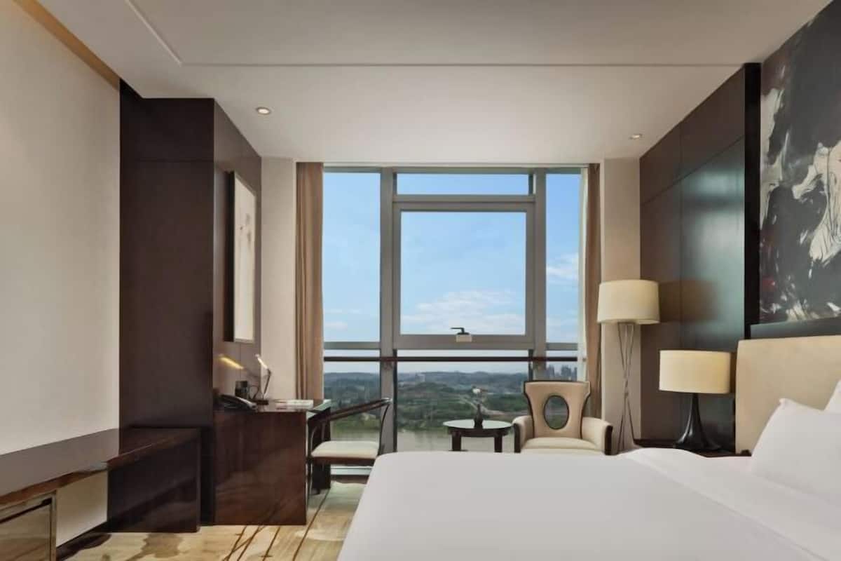A hotel room with a large window, a bed, a desk, a chair, and a lamp.