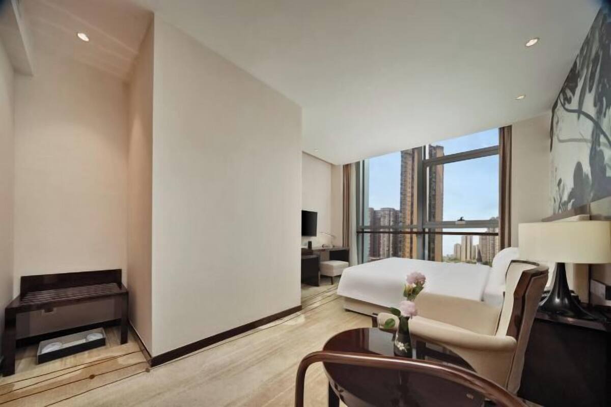 A modern hotel room with a large bed, a desk, a chair, a sofa, and a view of the city through large windows.