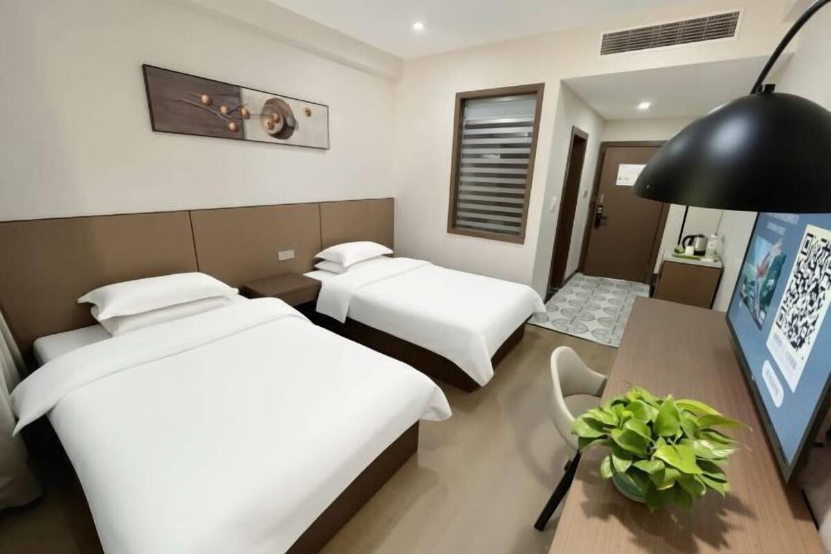Ein Hotelzimmer mit zwei Betten, einem Schreibtisch, einem Sessel, einer Zimmerpflanze, einem Fenster mit Jalousien, einer Tür und einem Wandbild.
