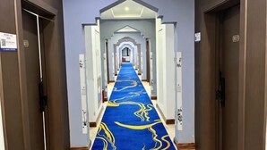 Een hotelgang met een blauw tapijt, meerdere deuren en een plafondlamp.