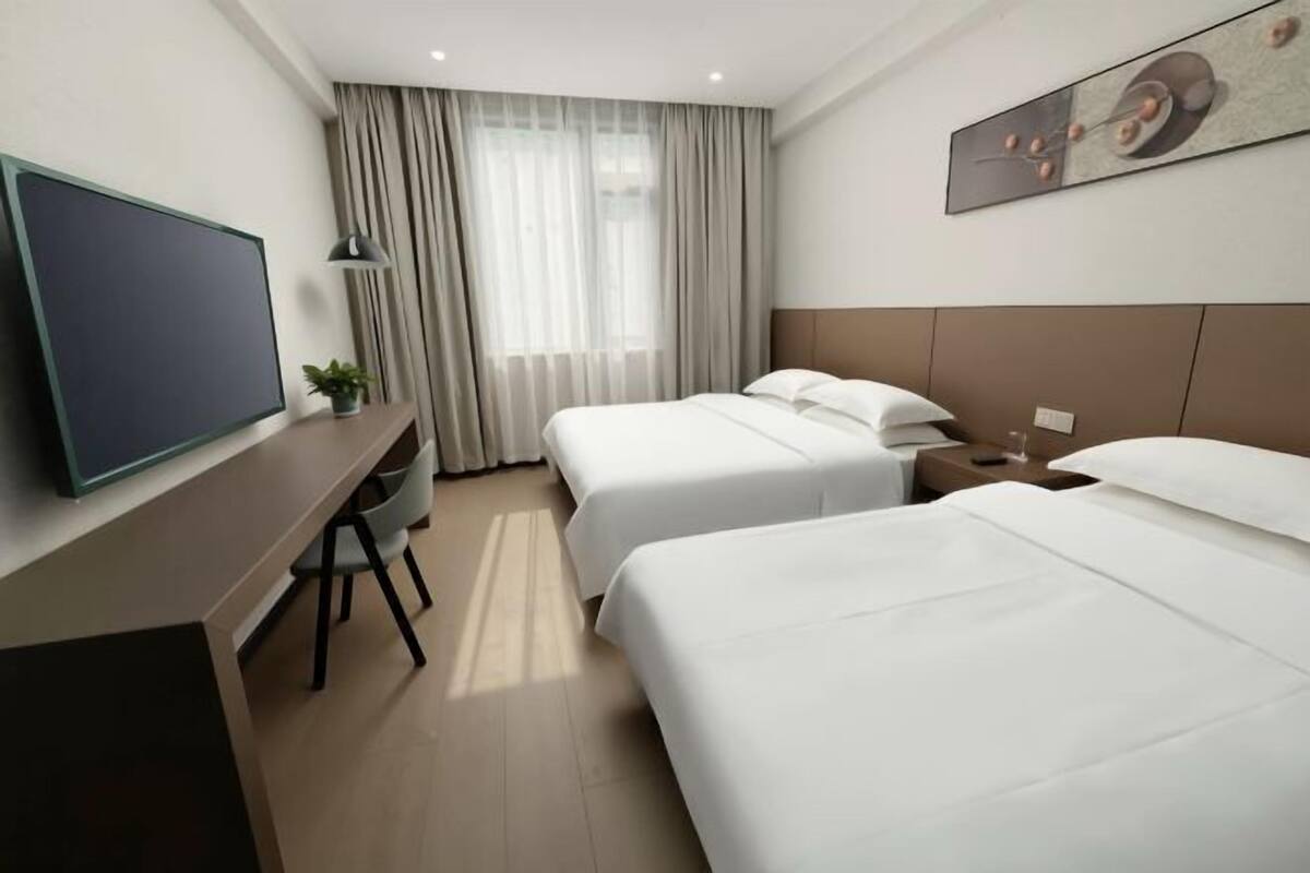 Ein Hotelzimmer mit zwei Betten, einem Schreibtisch mit Fernseher, einem Sessel und einem Fenster mit Vorhängen.