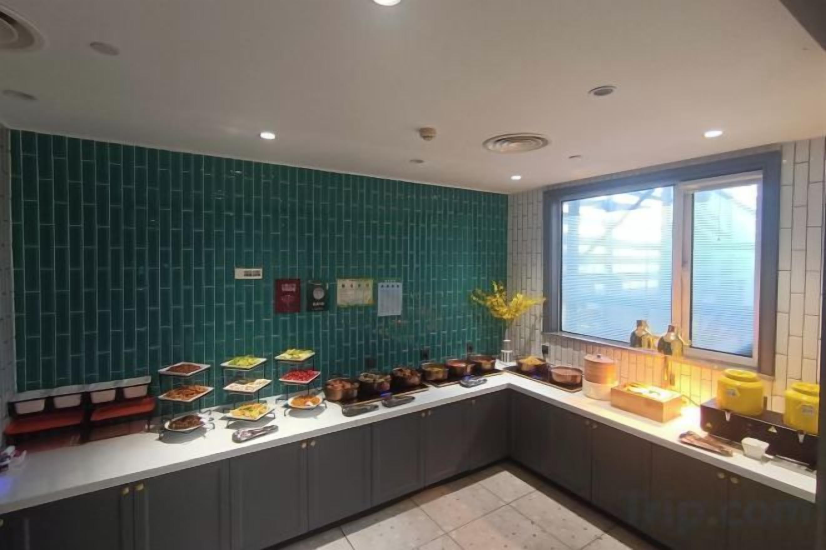 Una cocina moderna con salpicadero verde azulado, una encimera larga con diversos alimentos y un ventanal que proporciona luz natural.
