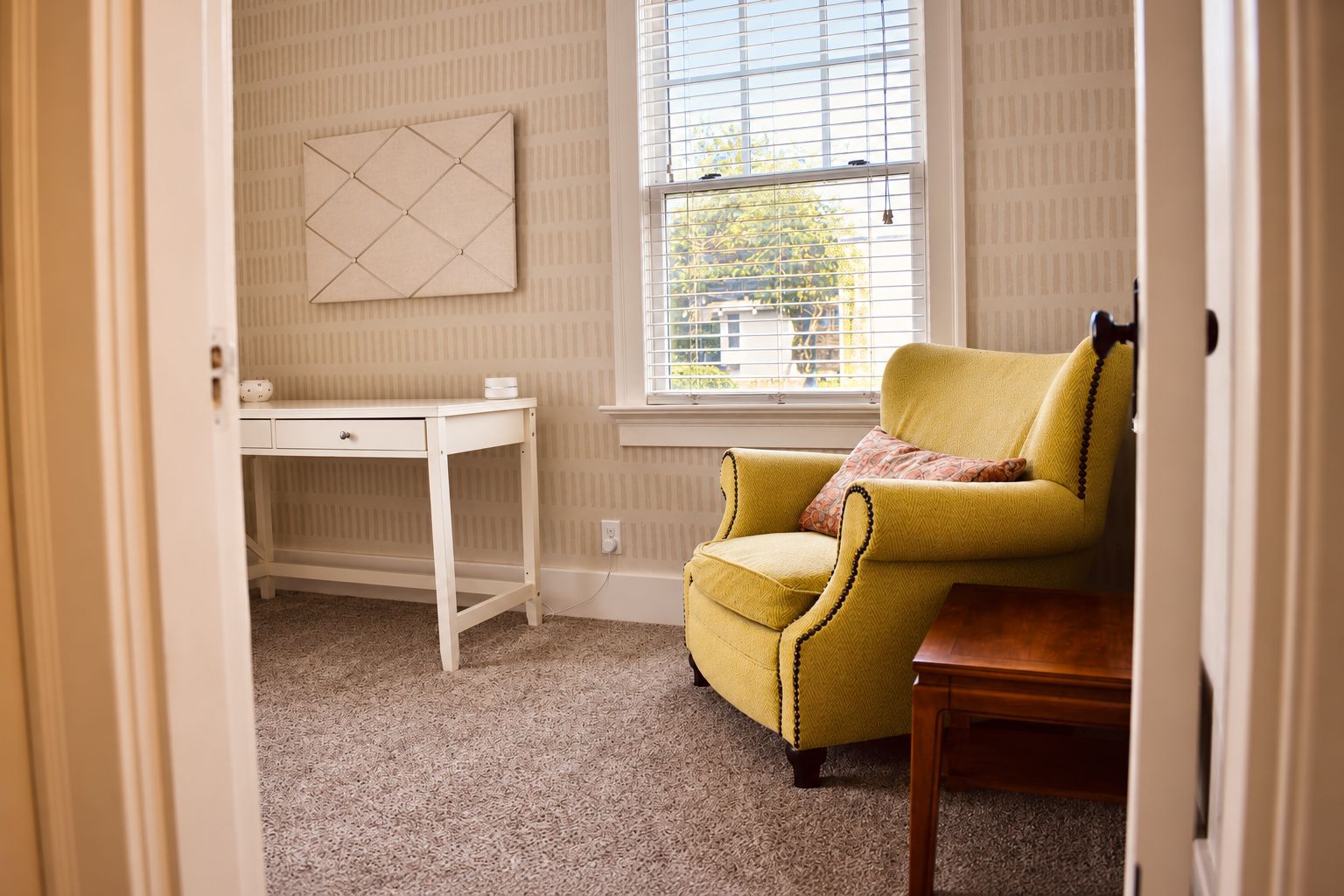 Una habitación con un sillón amarillo, un escritorio blanco y una ventana con persianas.